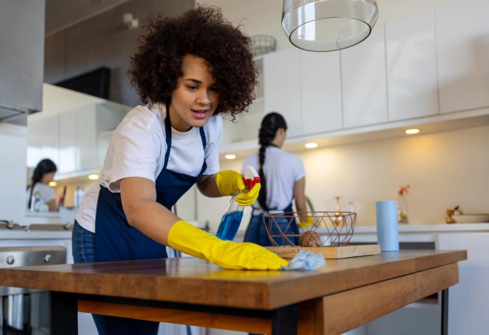 A woman in yellow gloves wipes down a table while others clean the kitchen behind her.