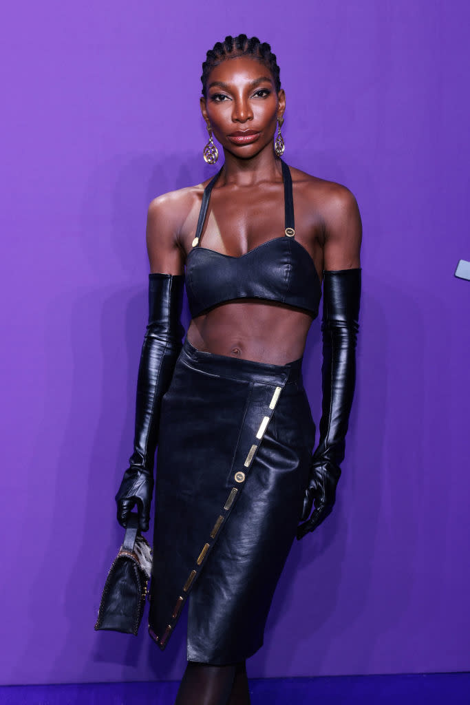 Michaela Coel on a red carpet, wearing a stylish leather crop top and skirt, long leather gloves, and holding a small clutch