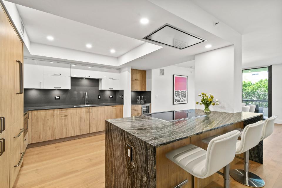 The kitchen has a combination of white high-gloss cabinets and high-end laminate cabinets. The waterfall-style island has a top and sides of textured “leathered” quartz.