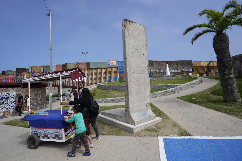 A family pushes a snack cart past a slab of the Berlin Wall, displayed near the border wall that separates the United States from Mexico, in Tijuana, Mexico, Friday, Aug. 25, 2023. The 3-ton pockmarked, gray concrete slab sits between a bullring, a lighthouse and the border wall, which extends into the Pacific Ocean. (AP Photo/Gregory Bull)