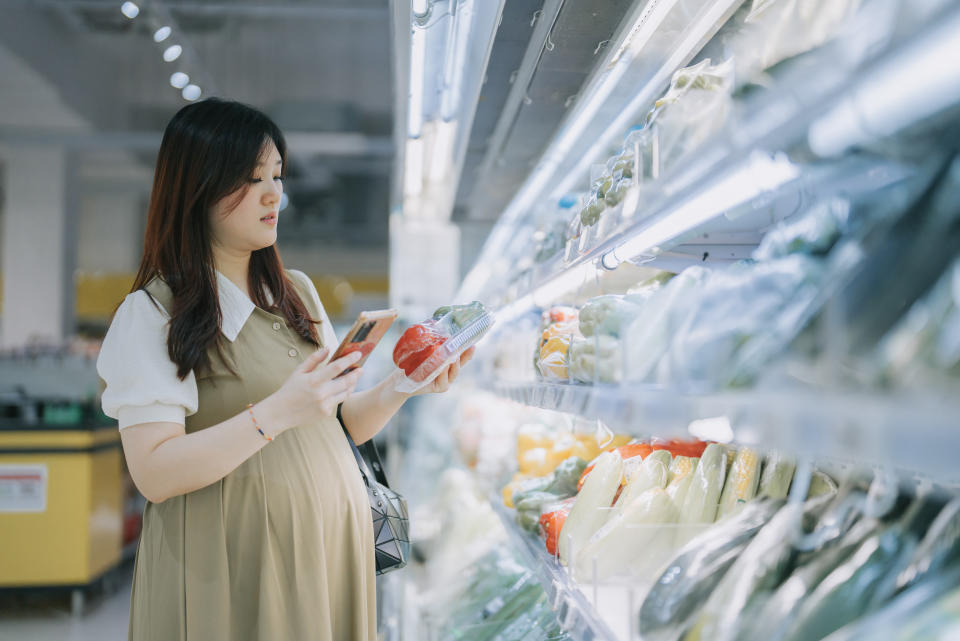 Asian Pregnant woman shopping buying vegetables  in supermarket refrigerated section surfing internet with smart phone