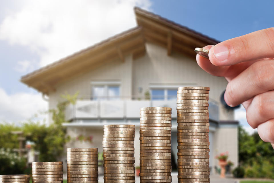 Human Hand Placing A Coin On Increasing Coin Stacks In Front Of House