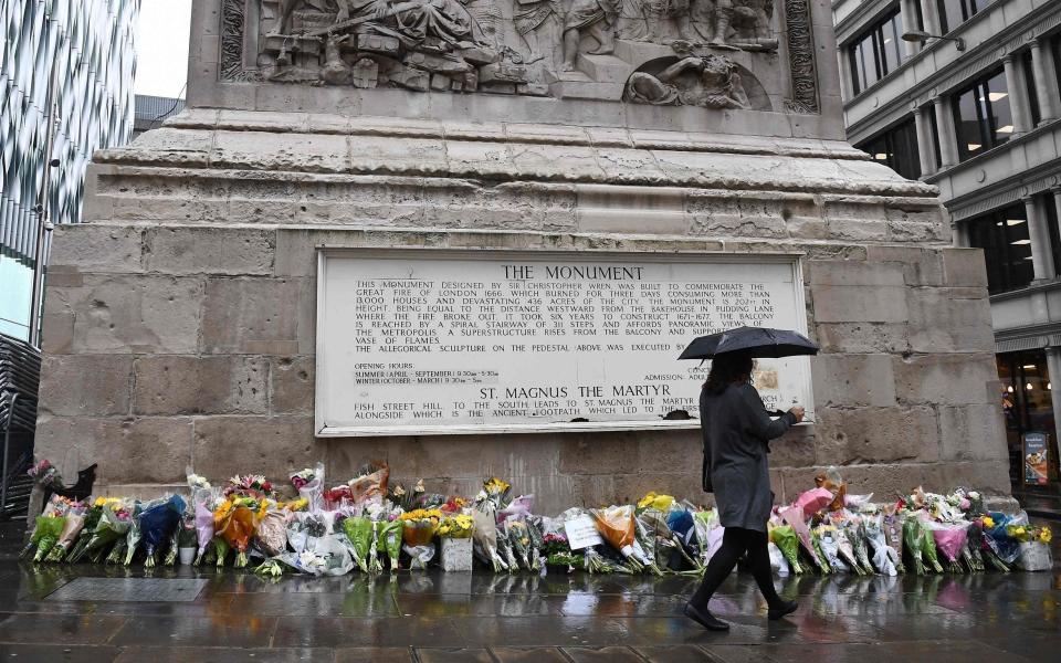 A commuter shelters from the rain beneath an umbrella as she passes floral tribute placed at the base of The Monument in London - Credit: JUSTIN TALLIS/AFP/Getty Images