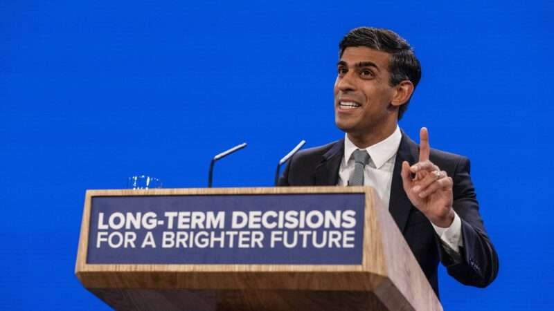 British Prime Minister Rishi Sunak speaking in front of a blue background, behind a podium that reads "LONG-TERM DECISIONS FOR A BRIGHTER FUTURE"