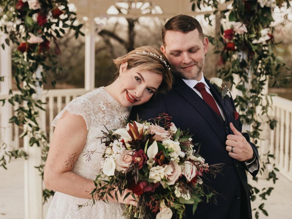 A bride leans her head on her groom's shoulder in front of a floral archway.