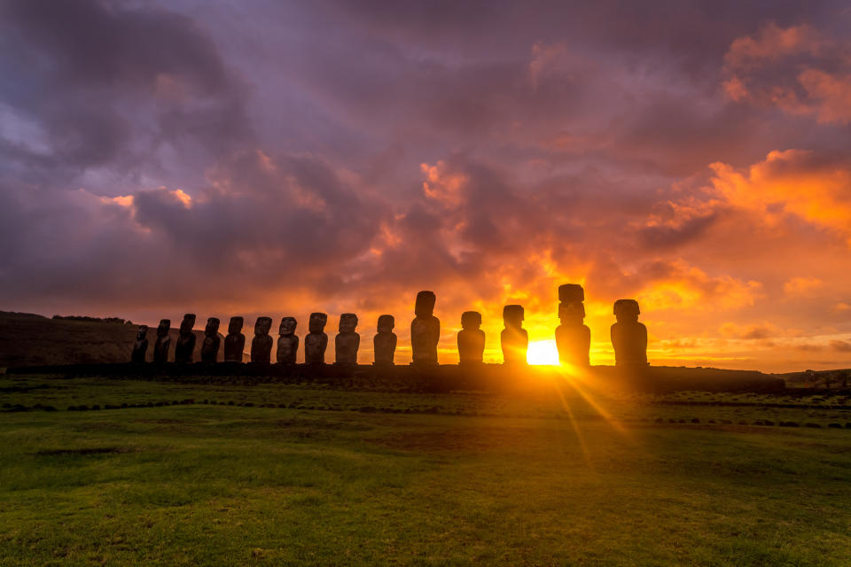 Several dozen tall stone-headed statues stand on a lush island under a colorful sunset