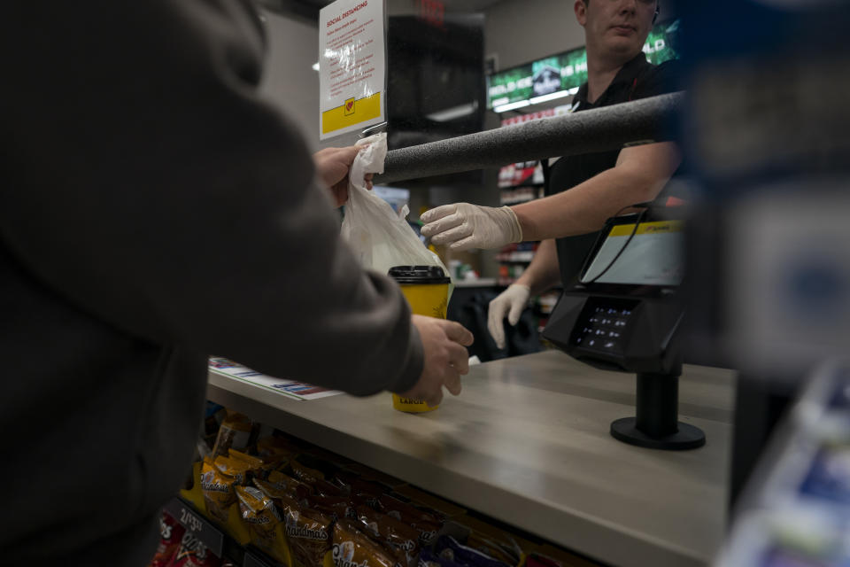 In this April 7, 2020, photo, a cashier behind protective glass wearing gloves hands trucker Sammy Lloyd, of Ringgold, Ga., his coffee and food at the Love's Travel Stop truck stop in Greenville, Va., before dawn. Lloyd was pulling a COVID-19 emergency relief load from California to a Target Distribution Center in Stuarts Draft, Va. (AP Photo/Carolyn Kaster)