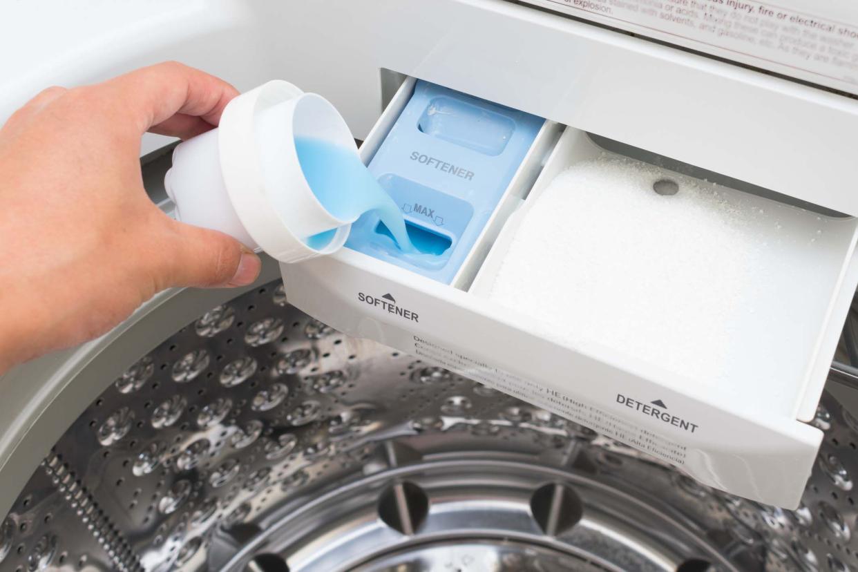 Hand holding cap pouring liquid softener into the detergent pan of a washing machine, light blue color, chrome washing basket in the background