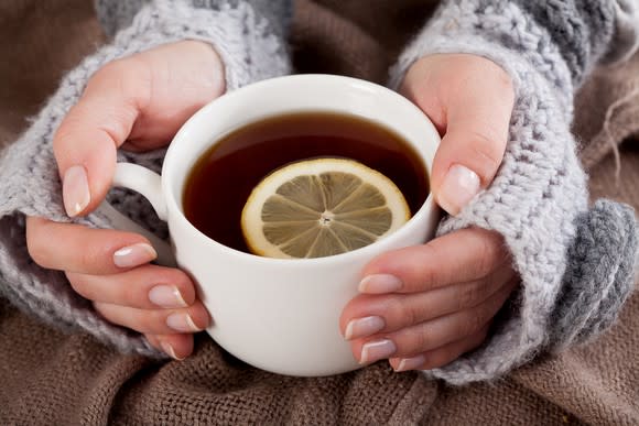 A lady's hands holding a cup of tea with a whole lemon slice in cup.