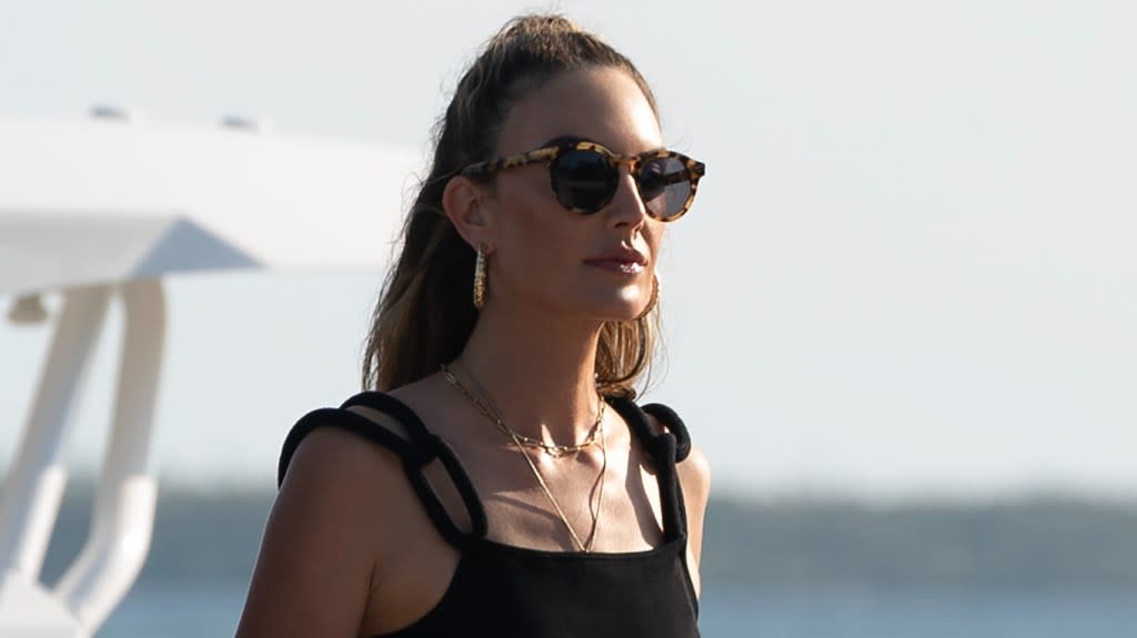 Elizabeth Chambers in a black tank top and black sunglasses walking on the beach