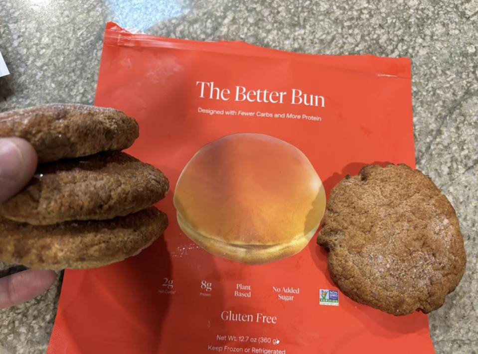 A hand holds two burger buns above a package labeled 'The Better Bun', highlighting the product's nutritional benefits