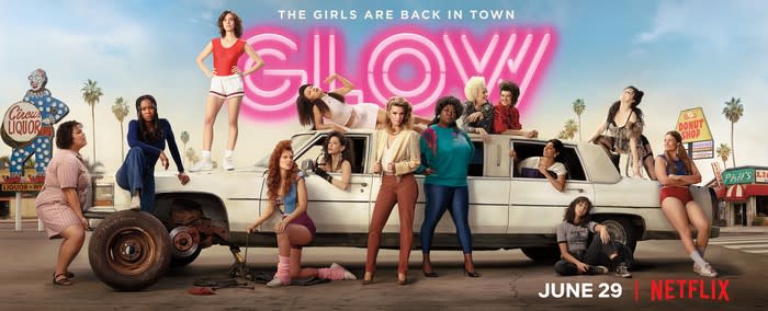 A bunch of women standing next to, leaning against, and standing or laying on a large car, with the caption The Girls Are Back in Town -- GLOW.