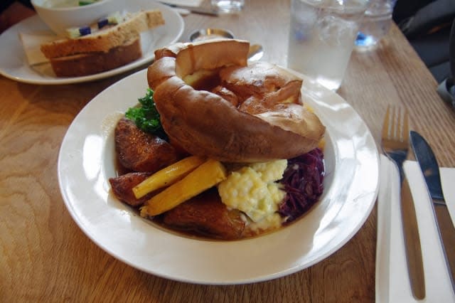 Roast vegetables, cauliflower cheese, and boiled red cabbage topped with a large Yorkshire pudding, served in a round white plat