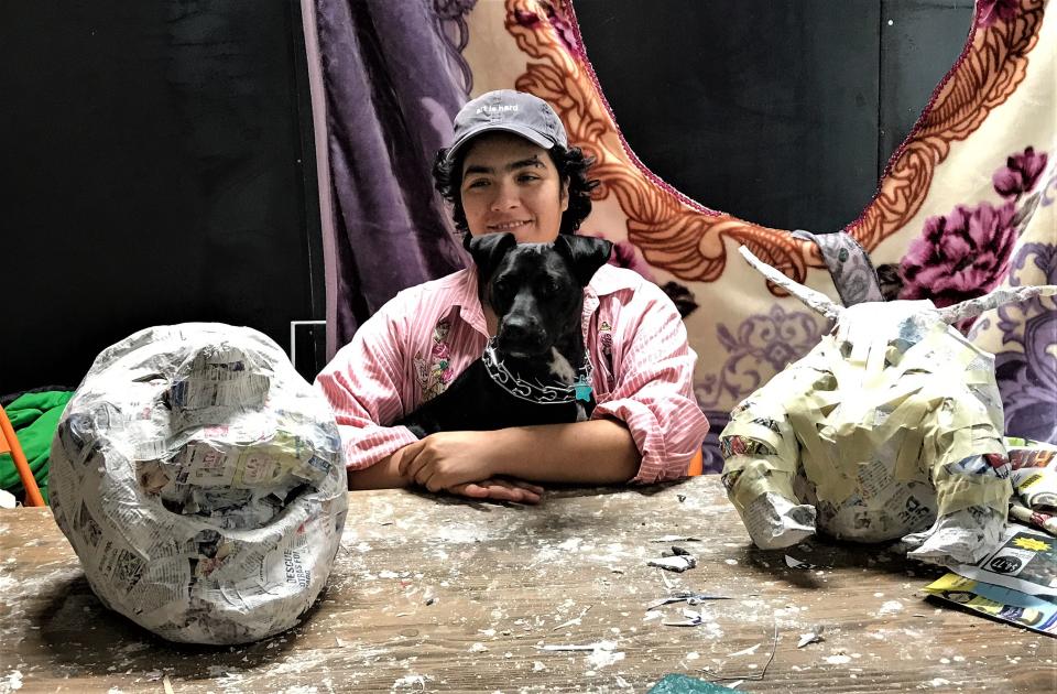 Bella Varela and her furry friend Darla are flanked by papier-mache works Varela is creating. A blanket she is using for her textile artwork is behind the two.
