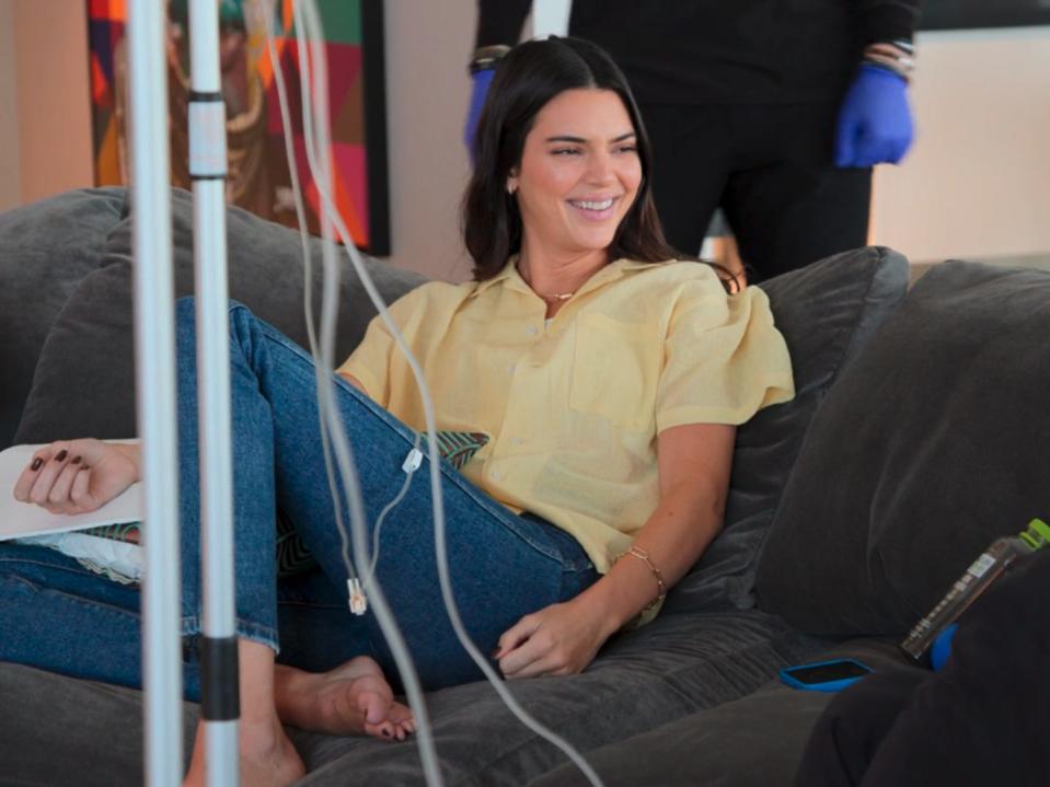 kendall jenner sitting casually on a couch in a yellow shirt and jeans, with an IV running into her arm. she's smiling