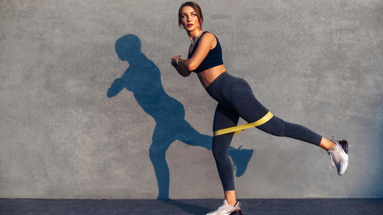  A woman performing a standing donkey kick with a resistance band around her legs. 