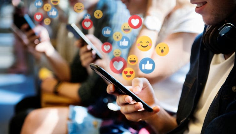 A group of young people staring at their phones with social media emojis floating up into the air.