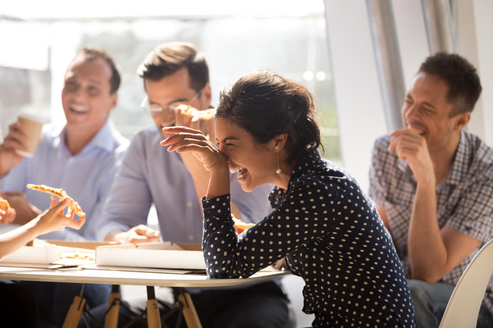 Indian woman laughing at funny joke eating pizza with diverse coworkers in office, friendly work team enjoying positive emotions and lunch together, happy colleagues staff group having fun at break