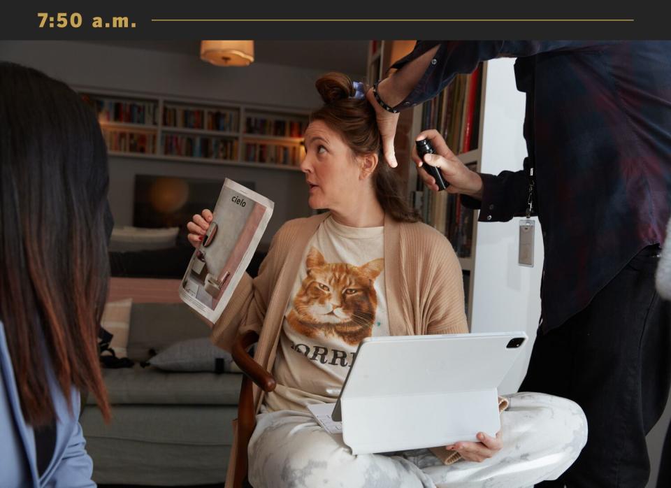 Drew Barrymore, with a tablet on her lap, is getting her hair done and holding up a paper to show someone.