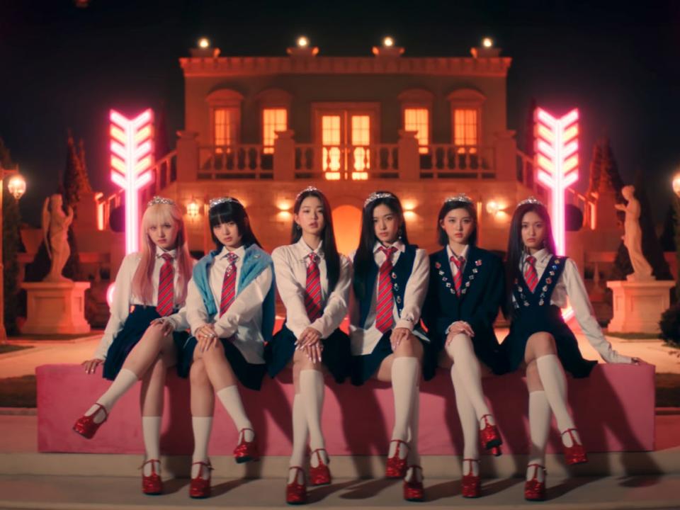 six young women sitting on a pink bench at night in front of a mansion. they're all wearing tiaras, sparkling pink shoes, and pink striped ties, with their legs crossed and serious expressions on their faces