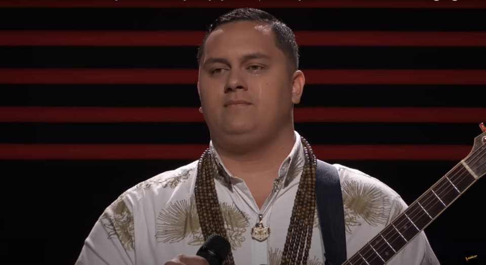 Kamalei stands with guitar in front of American flag background, appearing emotional