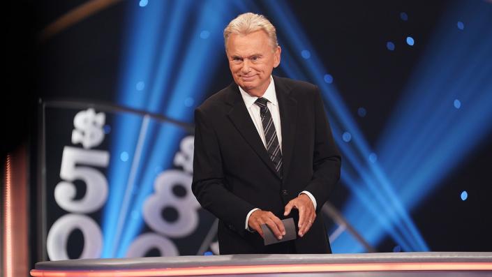 Pat Sajak stands behinds a barrier wearing a black suit and a patterned tie holding a card taping &quot;Celebrity Wheel of Fortune&quot;