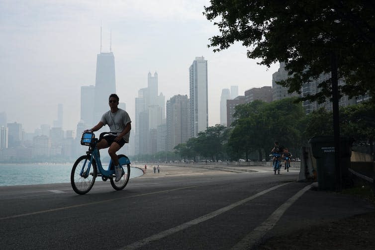 A young man rides a rental bike along Chicago's Lake Michigan shore with smoke obscuring the view of the city skyline in the background.