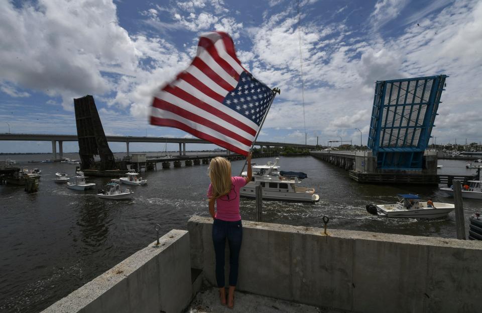 Suzanne Deuser, of Stuart, waves a flag in support of boaters as they travel through the open railroad bridge (left) and the Old Roosevelt draw bridge in the St. Lucie River on Monday, July 25, 2022, in Stuart. "Supporting a good cause," Deuser said while flying the flag in the breeze and cheering on the passing boats as they protest agains long railroad bridge closures to allow for Brightline train traffic.