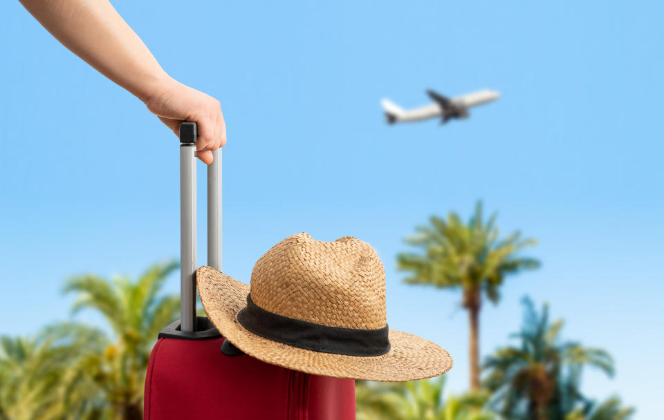 A hand pulling a suitcase with a straw hat on top, palm trees, and an airplane in the background, suggesting travel, possibly for a honeymoon