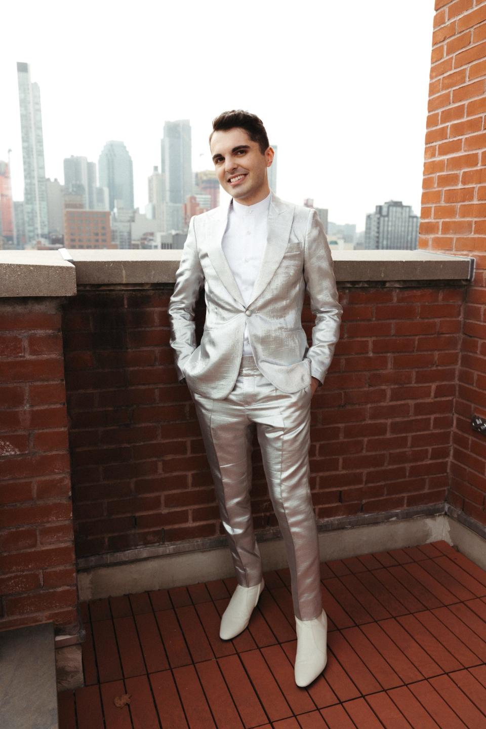 A man in a silver suit and white shoes stands on a brick balcony.