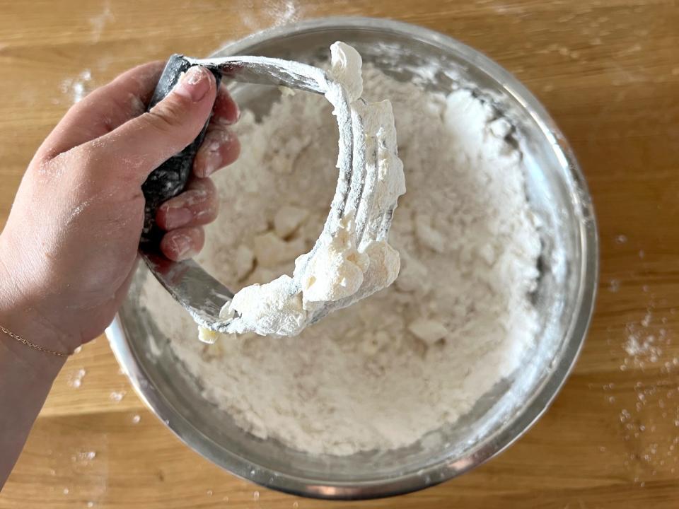 A hand holds a pastry cutter with butter stuck on it above a bowl of flour.