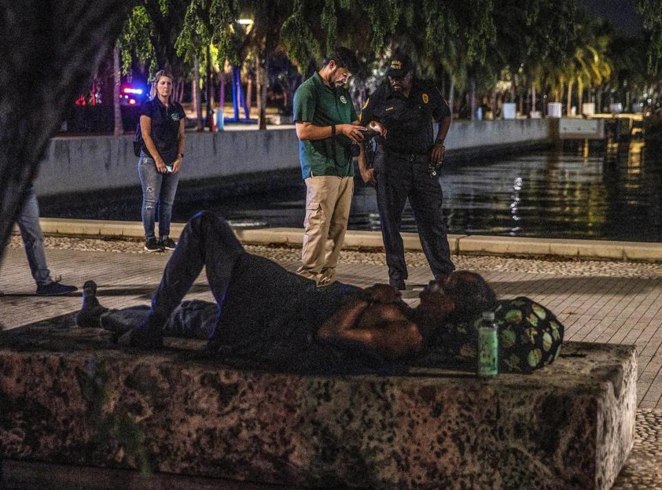 Un indigente duerme en un banco en Museum Park mientras los especialistas de Vinculación de Miami Christian Candelier (izquierda) y Ricky Leath caminan por el parque durante el censo anual de verano de personas sin hogar del Condado Miami-Dade, el jueves 18 de agosto de 2022.