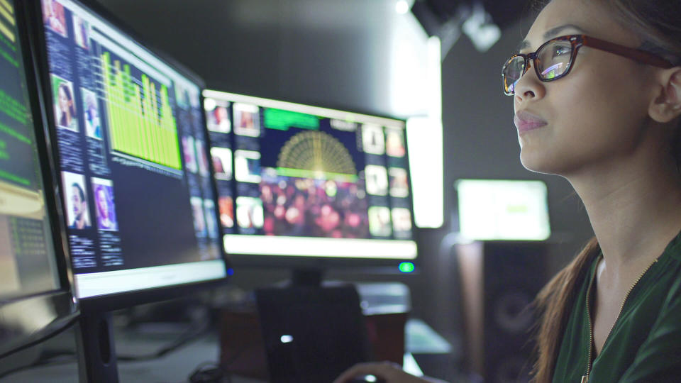 Close up stock image of a young asian woman sitting down at her desk where she’s surrounded by 3 large computer monitors displaying out of focus images of people as thumbnails; crowds; graphs & scrolling text.