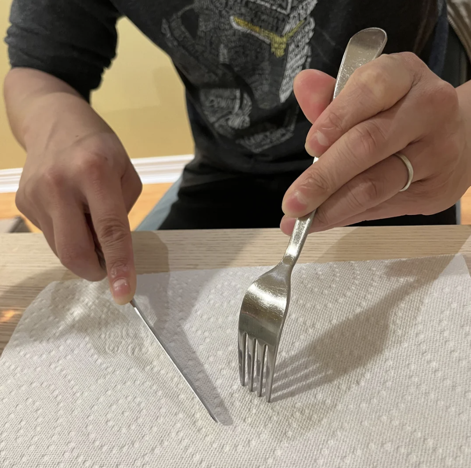 A person with a wedding ring is using a knife and fork on a table covered with a paper towel. The person's head is not visible in the image