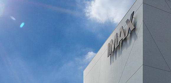 IMAX logo in the form of a sign at the corner of a tall building, set against blue skies.