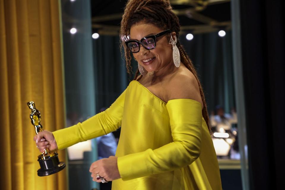 A smiling woman in a yellow dress holds an Oscar.