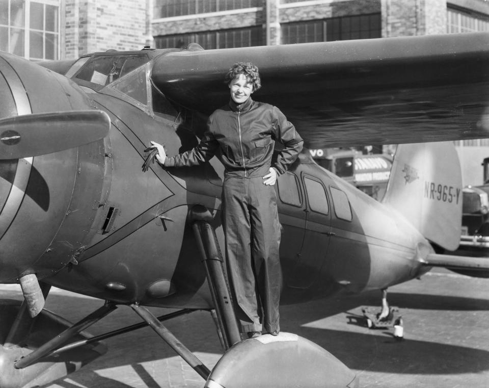 amelia earhart stands on the wheel of a plane, one hand rests on the aircraft and one hand is on her hip, she wears a jacket and pants