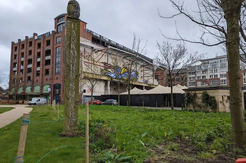 Bankmore Square off Belfast's Dublin Road, showing totem pole style wood post with name of square and grassy area with Trademarket and multi-story car park in background