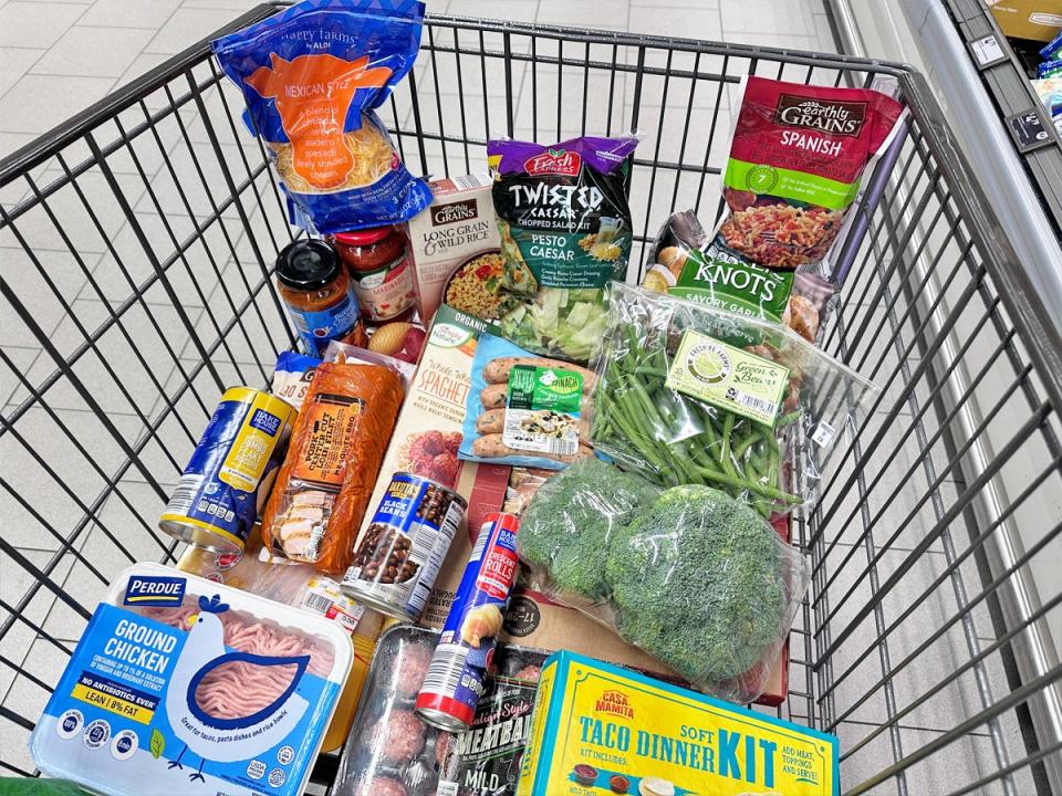 A shopping cart at Aldi full of broccoli, green beans, a yellow taco dinner kit, ground chicken with a blue design, canned items, a pack of sausages, and a blue and orange bag of shredded cheese