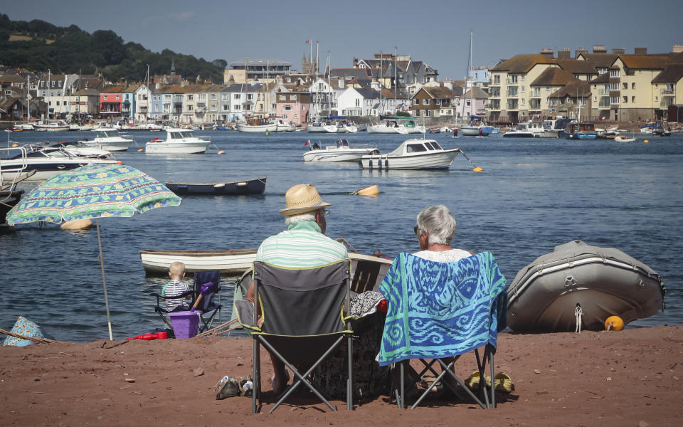 People sit on the beach facing the Teign estuary and the the holiday resort of Teignmouth in Shaldon, Devon, England, Wednesday July 21, 2021. Visiting the fishing village of Shaldon a small cluster of mainly Georgian houses and shops at the mouth of the River Teign, is like stepping back into a bygone era. It features simple pleasures that hark back to analog, unplugged summer days: a book and a picnic blanket, a bucket and spade, fish and chips. These are the traditional trappings of the great British seaside holiday that is making a comeback amid foreign travel concerns during the COVID-19 pandemic. (AP Photo/Tony Hicks)