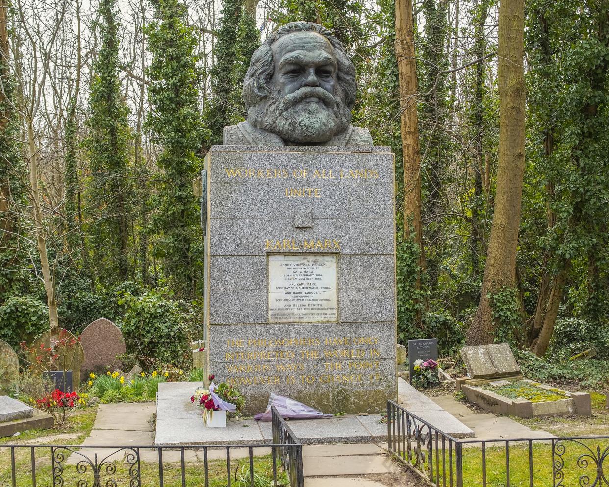 Karl Marx's tomb in the East Highgate Cemetery, London surrounded by a black fence and other smaller graves with the forest in the background on a winter