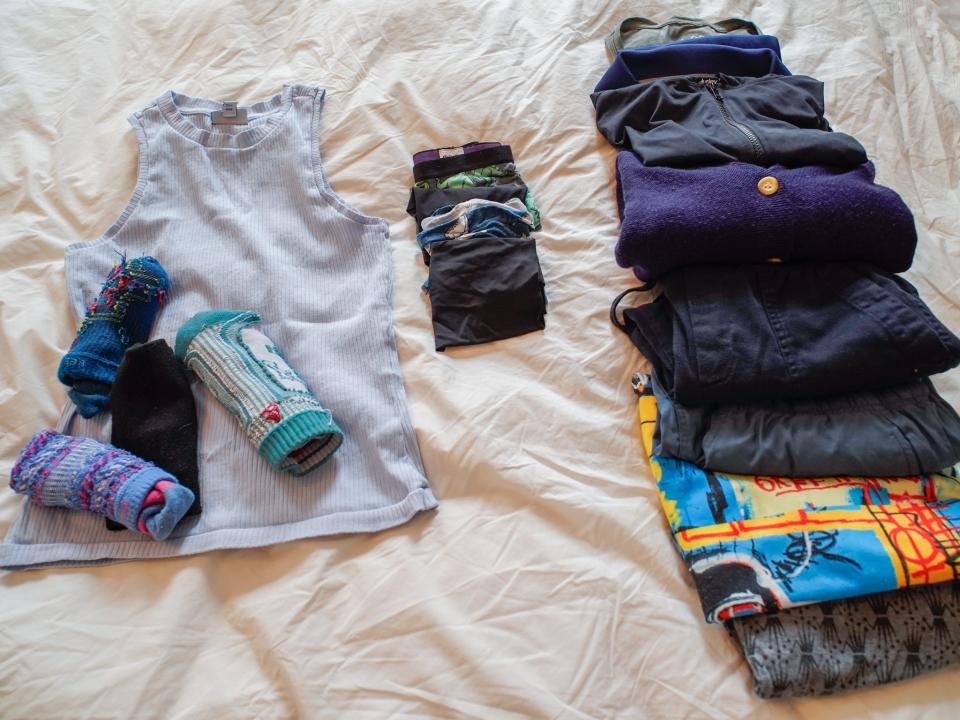 From left to right:  Four pairs of blue and black socks on a light blue tank top, five pairs of dark-colored underwear, and and a row of two rolled-up shirts and two fold-up long sleeve layers that are gray, black, navy blue folded teal pants, folded black pants, a folded blue and yellow t shirt, and folded gray leggings with a black pattern   on a white sheet