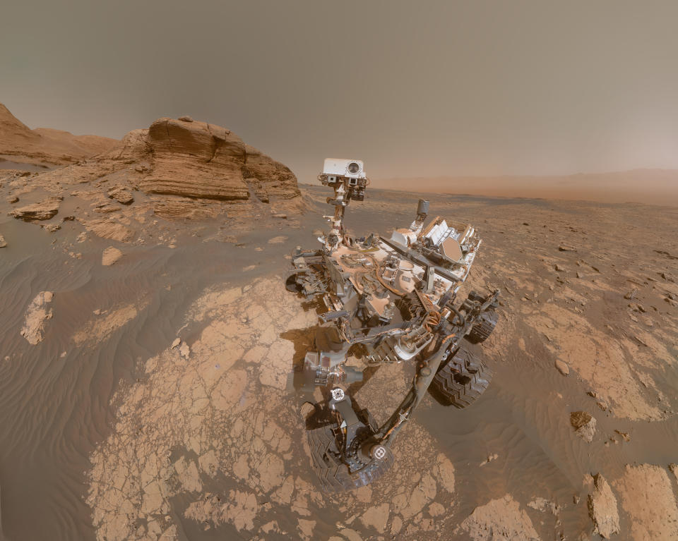 Selfie del rover Curiosity sobre la superficie de Marte, cerca de una pared rocosa llamada Mont Mercou, en el borde del cráter Gale, que se ve al fondo. (Imagen: Wikimedia Commons)