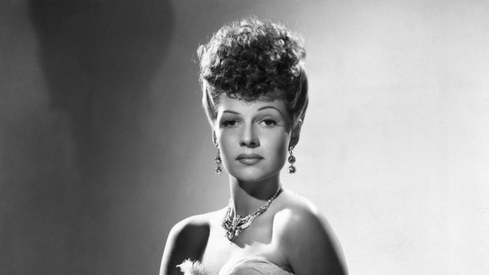rita hayworth sits on a cushioned stool and looks at the camera, she wears a strapless glittery gown with a necklace and dangling earrings