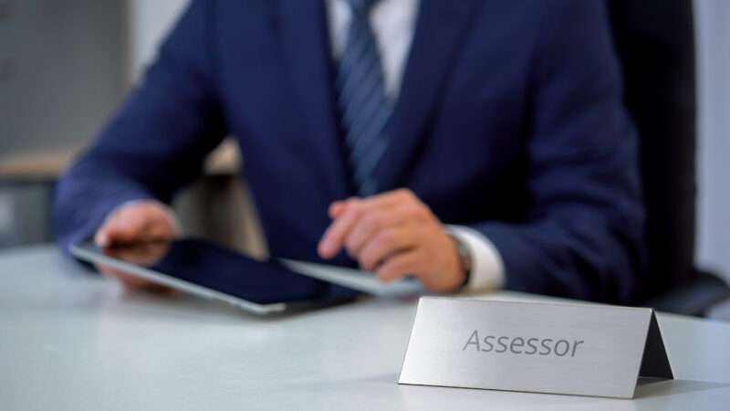 An unseen man in a business suit types on a tablet, at a desk with a nameplate that reads "Assessor"