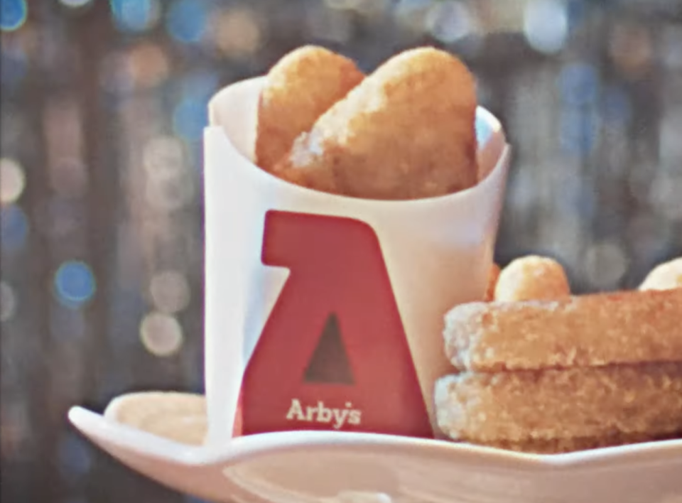 Arby's food tray with two mozzarella sticks in a branded container and several onion rings beside it. The setting appears festive with blurred lights