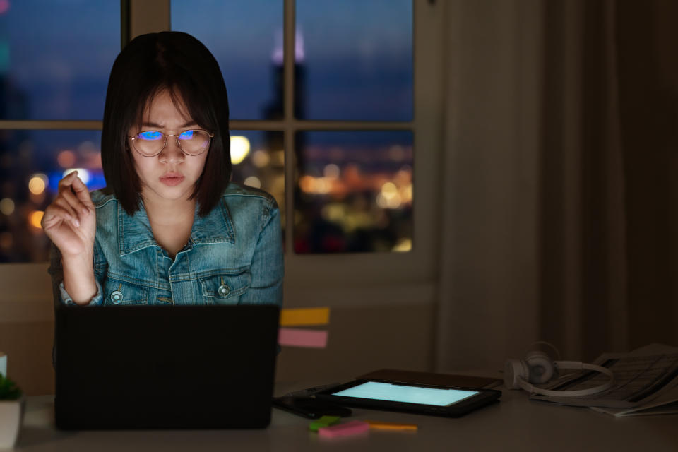 Candid of young attractive asian female student sitting on desk with smart digital gadget looking at notebook working at late night with project research, graphic designer or programmer concept.