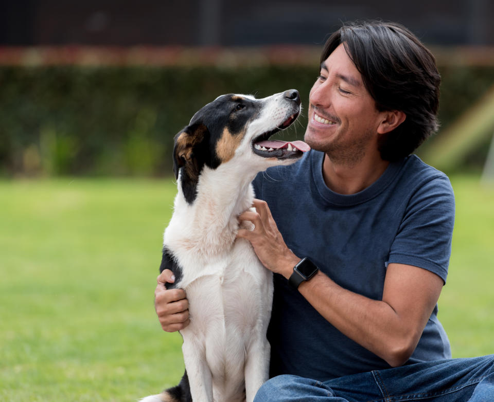 A man sits on grass and smiles at a dog, petting it gently while the dog looks back at him