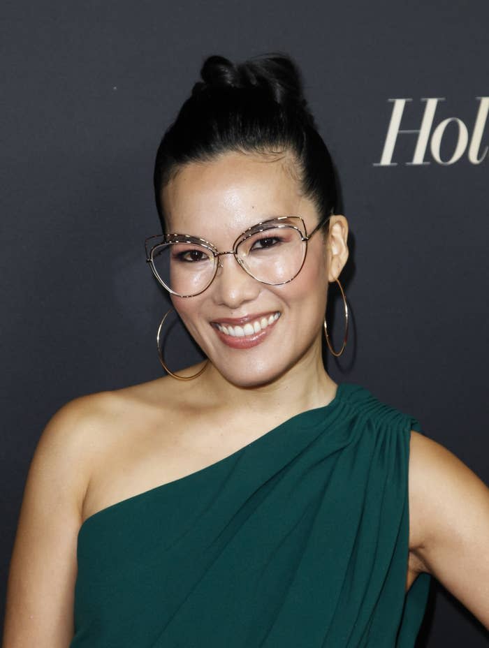 Ali Wong smiles for a photo at an event. She's wearing a one-shouldered outfit that ruched at the top of the shoulder and large hoop earrings. Her hair is swept into an updo