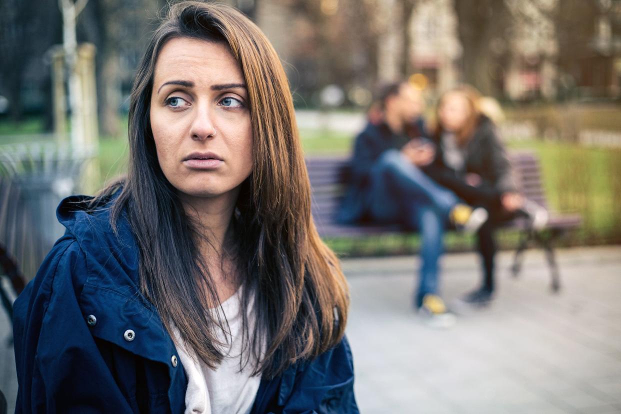 Moody woman sitting alone on a park bench on the left in the foreground, her eyes are looking towards the left, wearing a coat, a couple sitting at another park bench and park during winter on a sunny day, blurred in the background